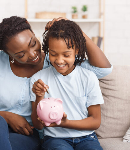 Little black girl putting money to piggy bank African Mother And Daughter Putting Coins Into Piggy Bank At Home. Free space