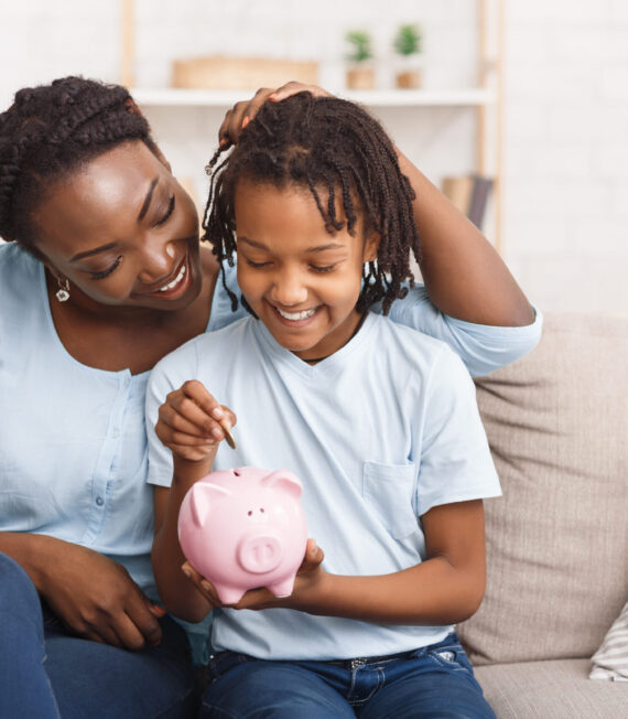 Little black girl putting money to piggy bank African Mother And Daughter Putting Coins Into Piggy Bank At Home. Free space