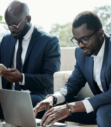 Young broker in elegant suit pressing keys of laptop keypad during network or while analyzing online financial data against colleague