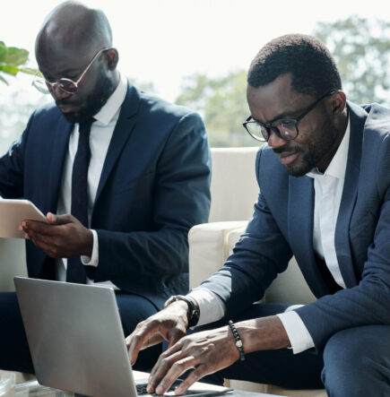 Young broker in elegant suit pressing keys of laptop keypad during network or while analyzing online financial data against colleague